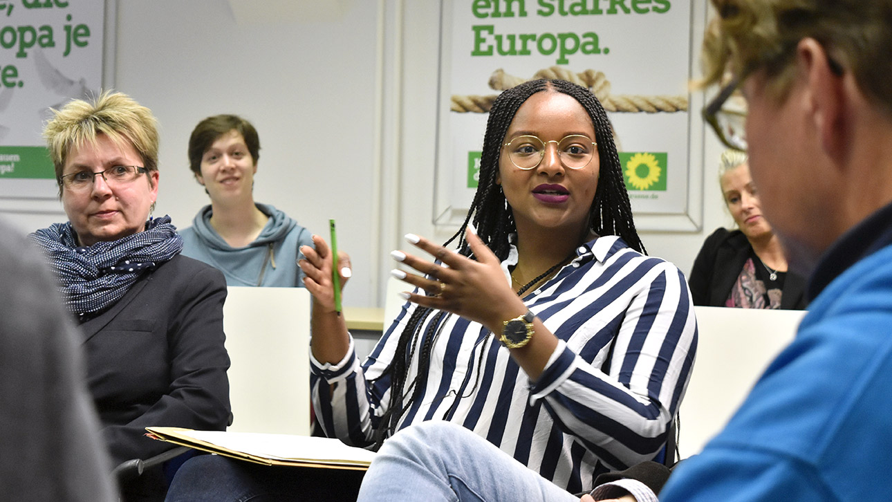 Die Grünen-Abgeordnete Aminata Toure sitzt zwischen mehreren Menschen in einem Saal mit Wahlplaketen der Grünen an der Wand. 