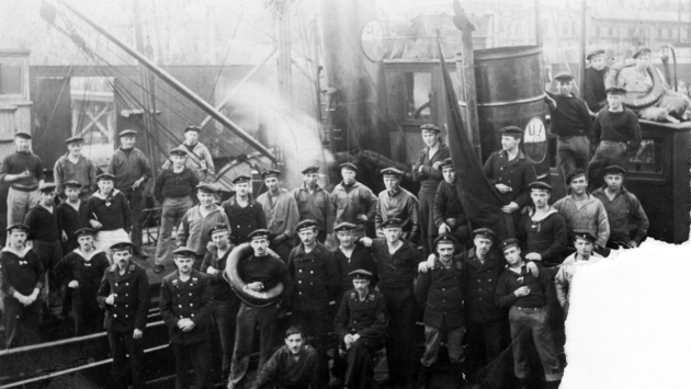 Auf einem Schwarzweißfoto vom November 2018 stehen Matrosen mit Fahnen und in Siegerposen vor einem Schiff, dem Minenräumboot Cladow