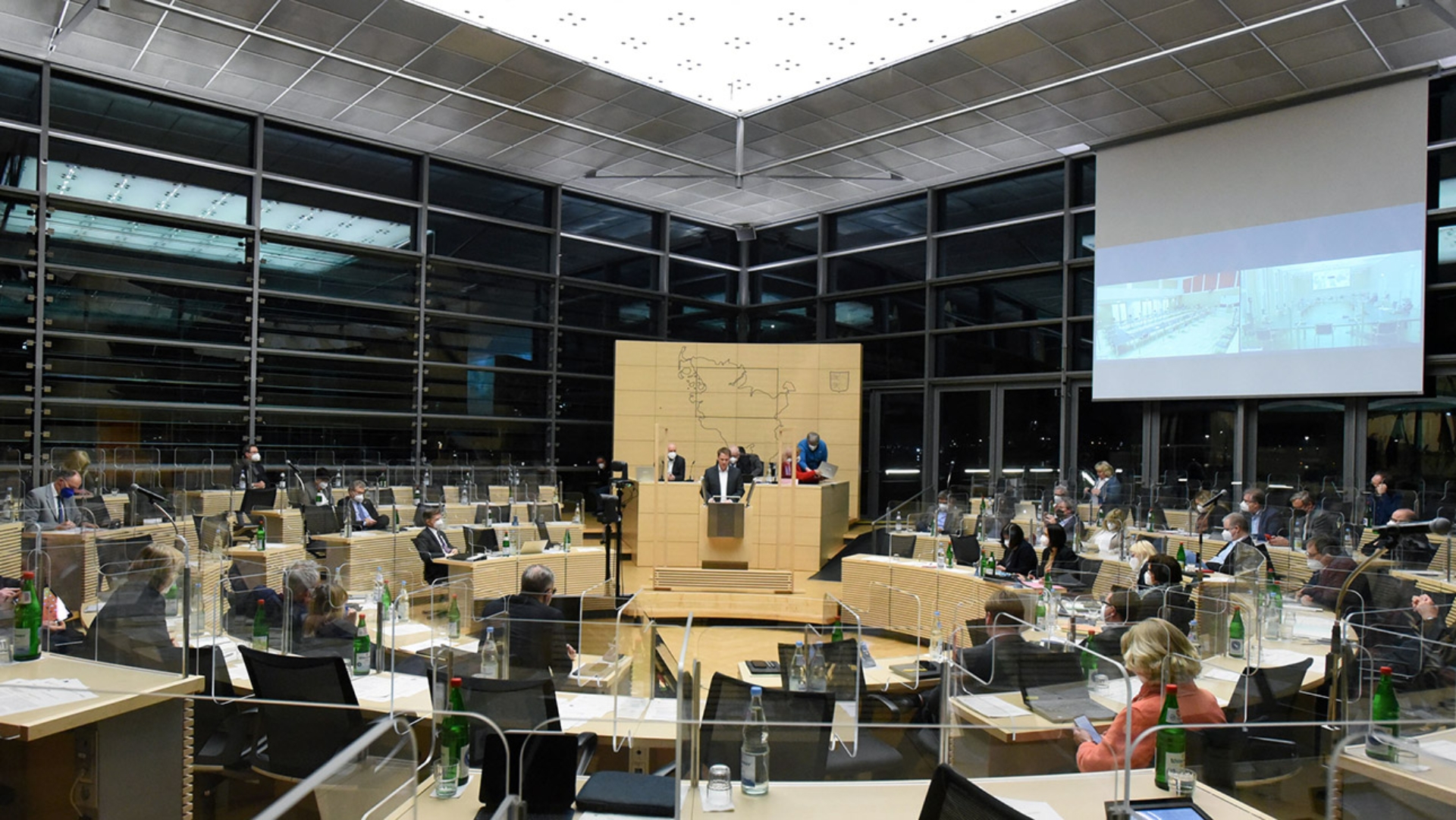 Blick in den mit Abgeordneten besetzten Plenarsaal, wo an einer Leinwand auch zwei weitere Räume mit Politikern eingeblendet sind.