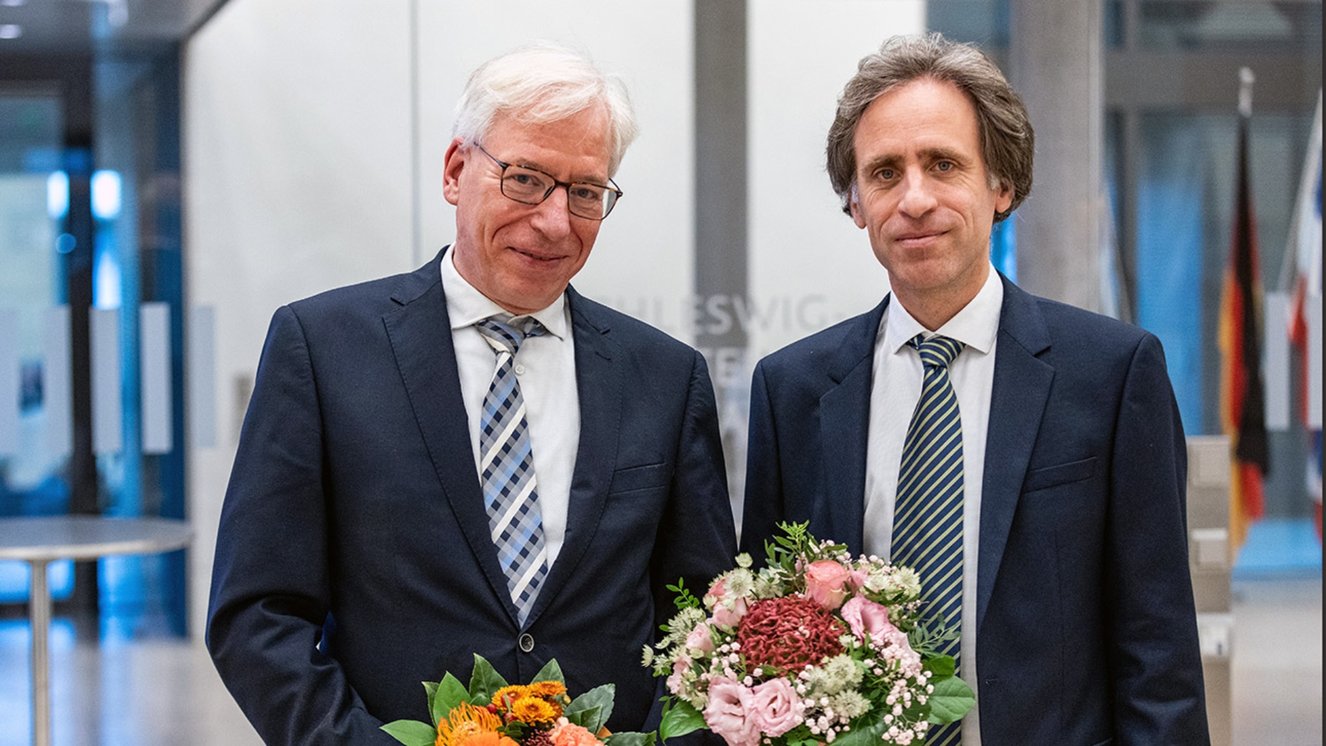 Pressefoto Carsten Löbbert und Dr. Marc Petit, die beide einen Blumenstrauss in den Händen halten.