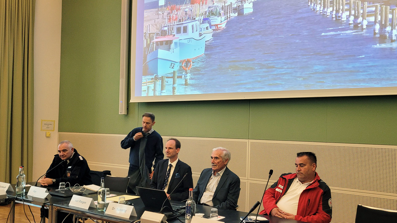 Mehrere Personen, darunter Petent Herbert Schmitz, Seenotretter Björn Hagge und Tilo von Riegen, Leiter der Kommunalabteilung im Innenministerium, sitzen vor einer Projektion, auf der der Hafen Lippe zu sehen ist.