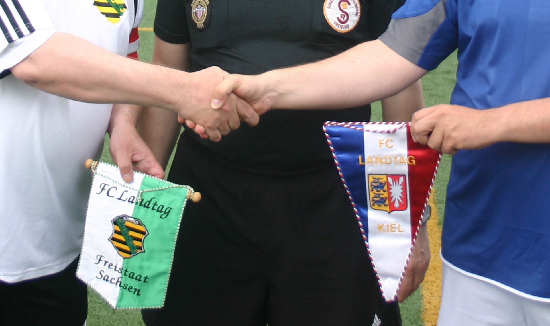 Zu sehen ist der von einem Händeschütteln begleitete Austausch von Wimpeln des FC Landtages und des FC Sachsen