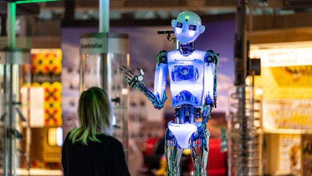 Eine Frau steht in einer Ausstellung vor einem bunt-strahlenden Roboter in Menschengröße. 