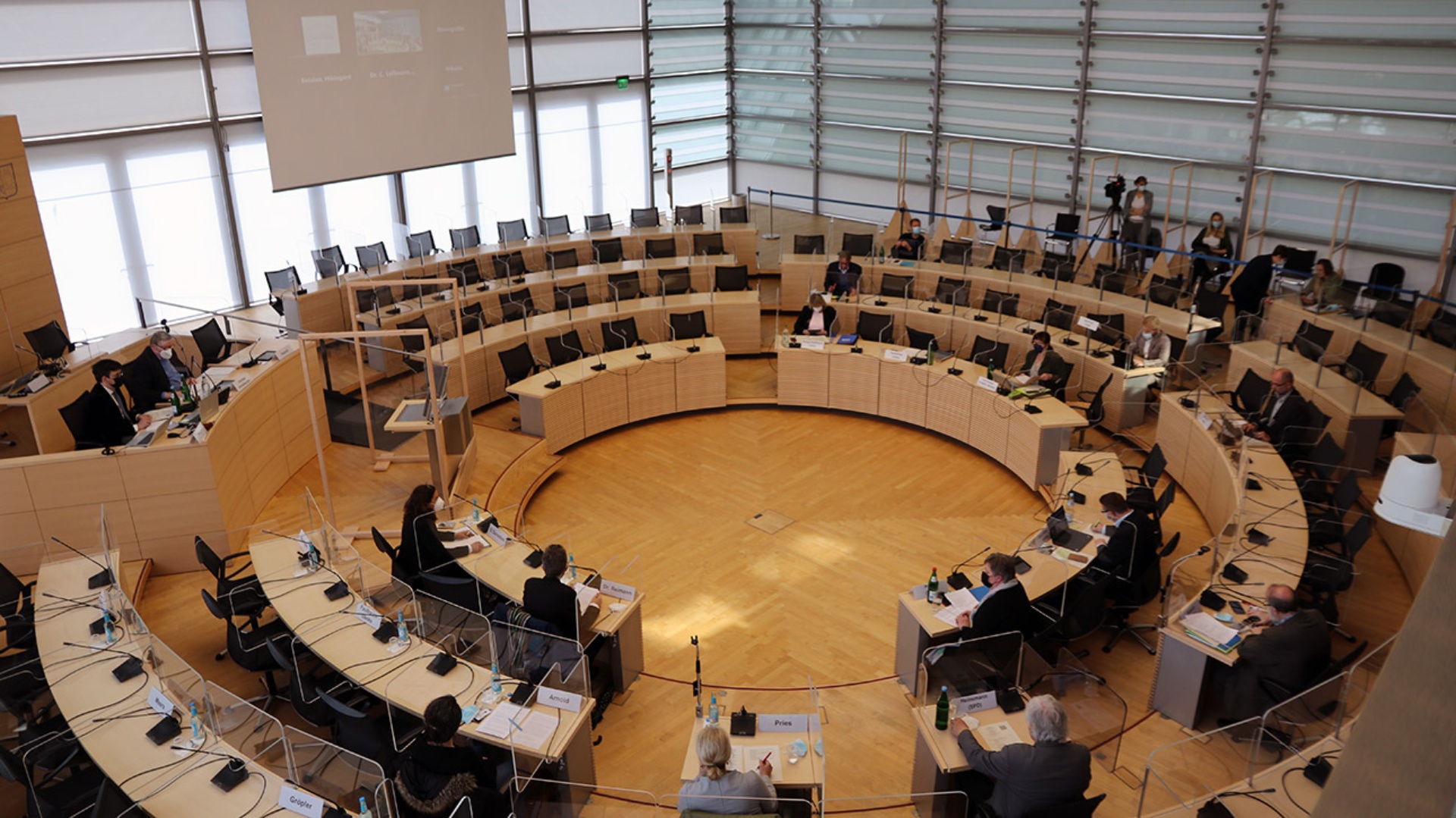 Blick von der Tribüne in den Plenarsaal, wo Abgeordnete, Anzuhörende und Kameraleute anwesend sind. An der Wand ist eine Leinwand mit der Projektion eines zugeschalteten Experten. 