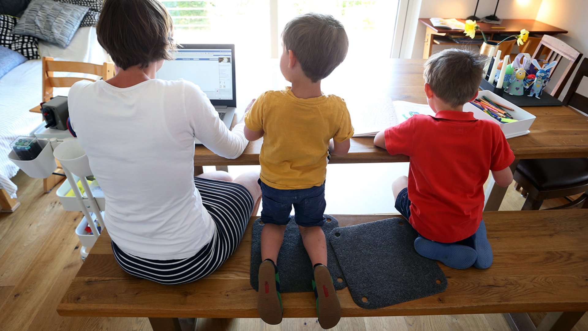 Die Mutter zweier Kinder arbeitet Zuhause an einem Laptop, während ihre Kinder neben ihr malen und ein Buch ansehen.