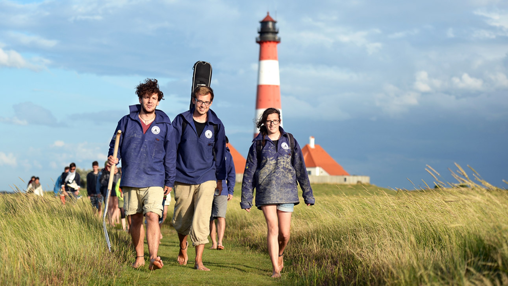 Drei junge Leute von der Schutzstation Wattenmeer führen eine Gruppe Wattwanderer an. Im Hintergrund ist der Leuchtturm in Westerhever.