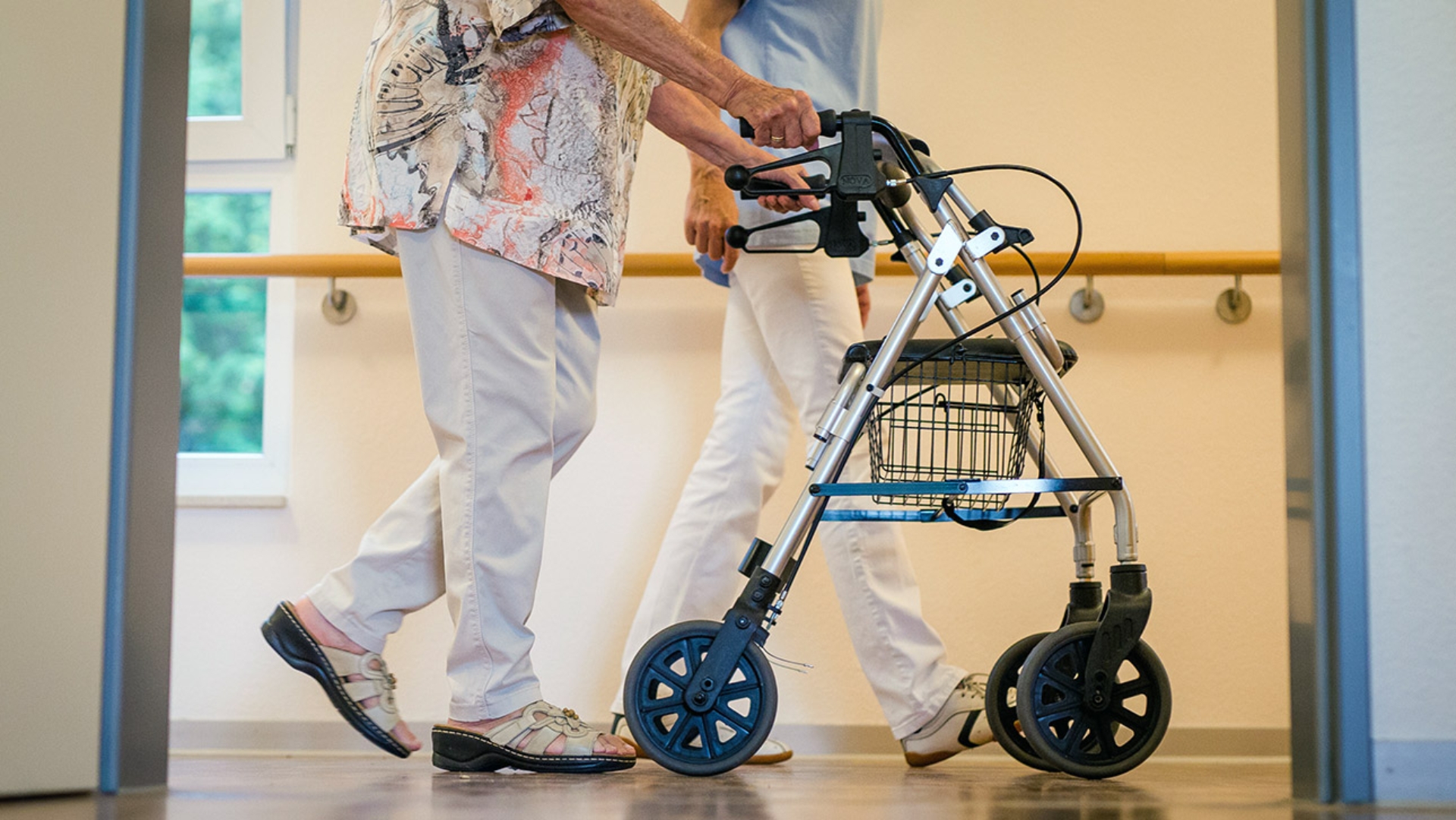 Eine Pflegekraft geht in einem Pflegeheim mit einer älteren Dame über einen Korridor. 