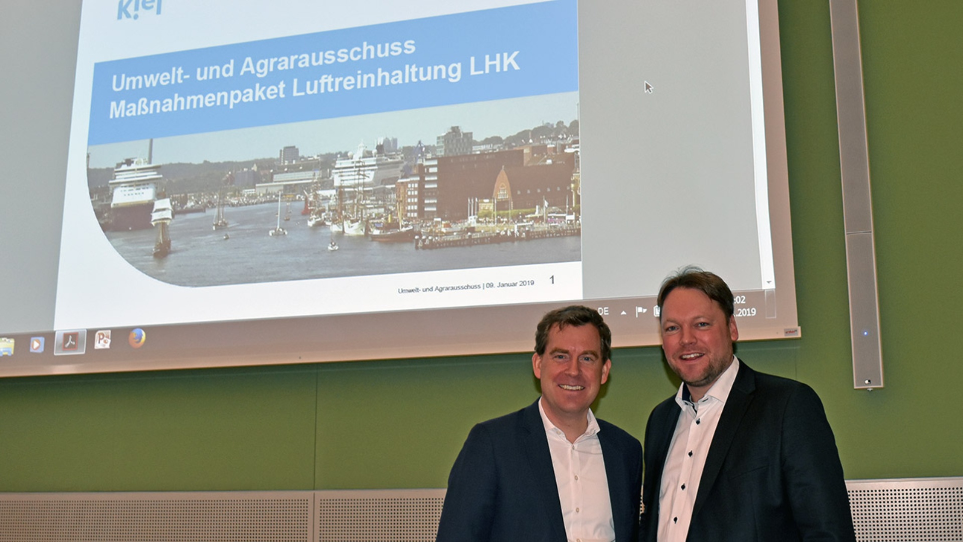 Der Ausschussvorsitzende Oliver Kumbartzky begrüßt Kiels Oberbürgermeister Ulf Kämpfer im Ausschusssaal des Landeshauses. Im Hintergrund ist eine große Projektion des Titelbildes des Luftreinhaltekonzepts der Stadt Kiel zu sehen.