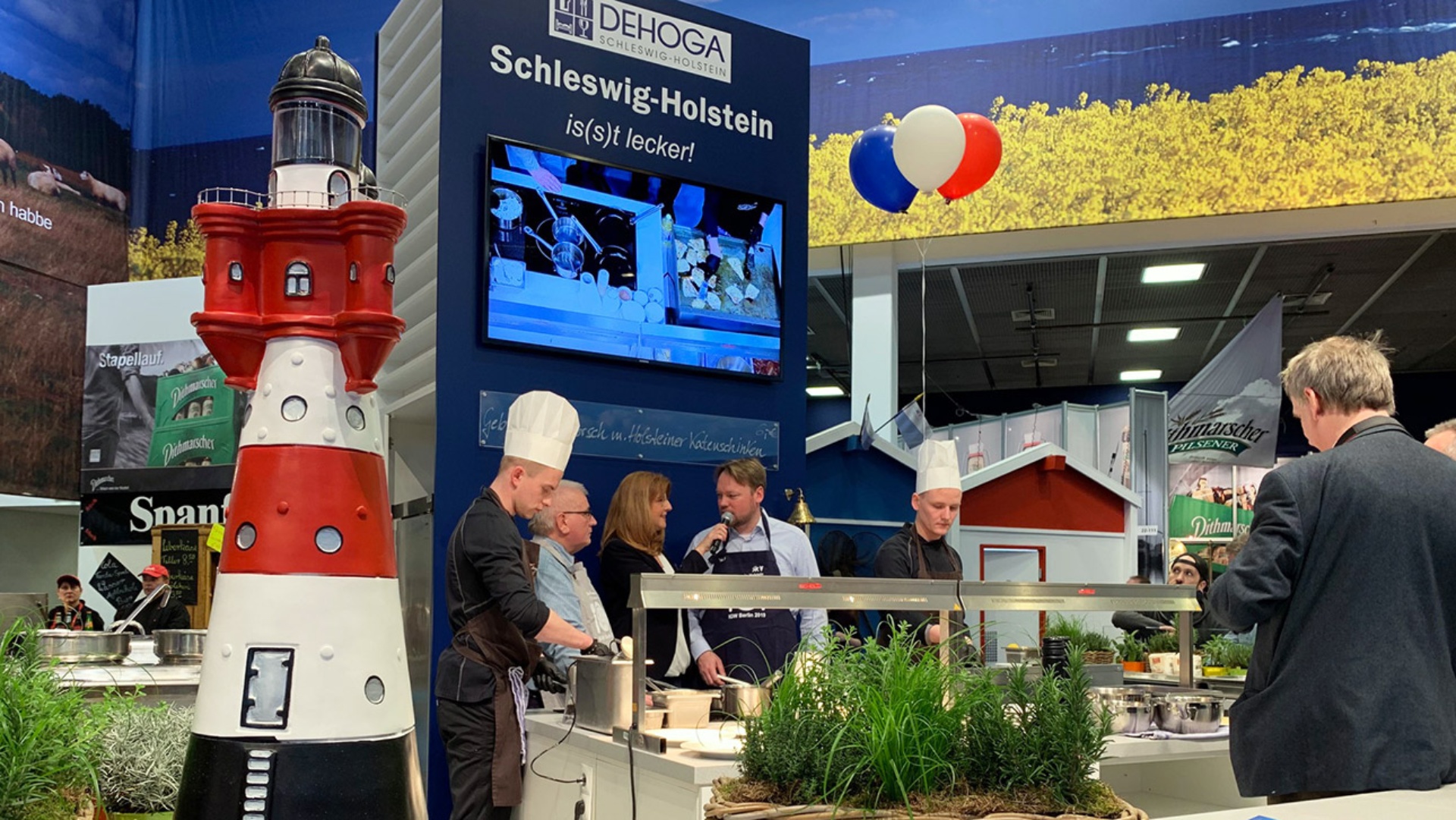 Landtagspräsident Schlie und der Agrarausschussvorsitzende Kumbartzky stehen mit zwei Köchen am Stand des Hotel- und Gaststättenverbands Schleswig-Holstein und werden interviewt. Im Vordergrund ein rot-weißer Leuchtturm.