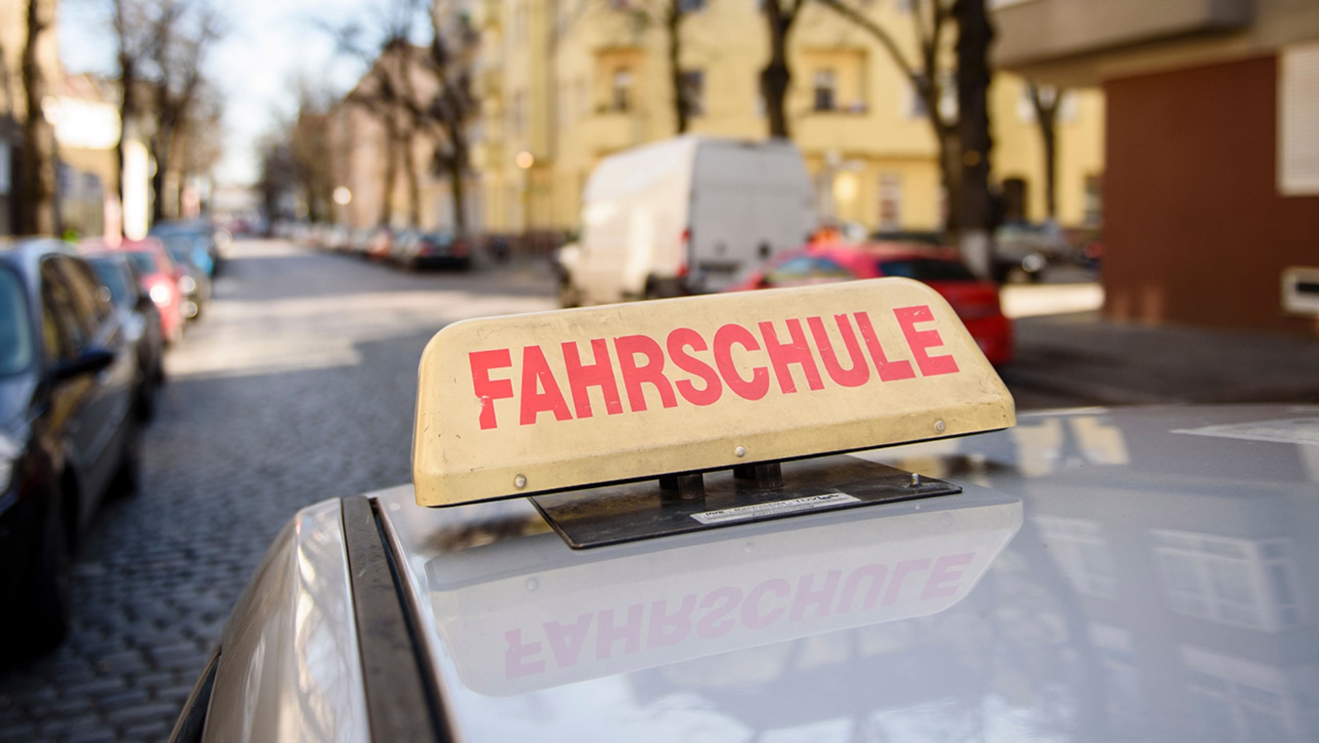 Auf dem Dach eines silbernen Autos ist ein Schild mit der Aufschrift „Fahrschule“ zu sehen.