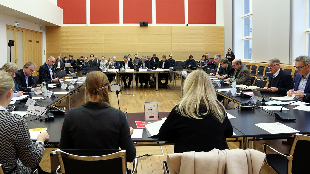 Blick von der Ausschussvorsitzenden in den Ausschusssaal: Rechts und links an Tischen sitzen Politiker, hinten drei Anzuhörende sowie rund 20 Zuhörer.