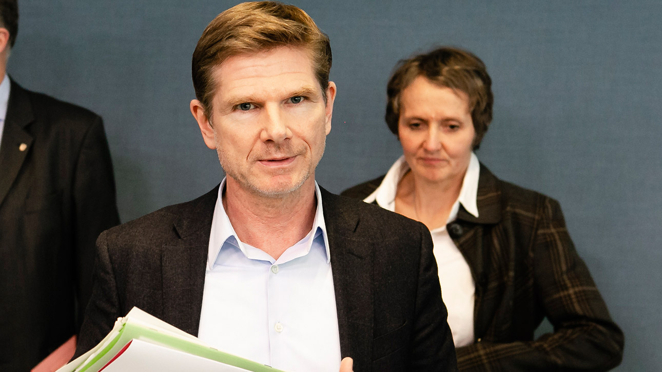 Sozialminister Heiner Garg hält einen Stapel Unterlagen in der Hand. Hinter ihm steht die Infektionsschutzreferentin des Ministeriums, Anne Marcic.