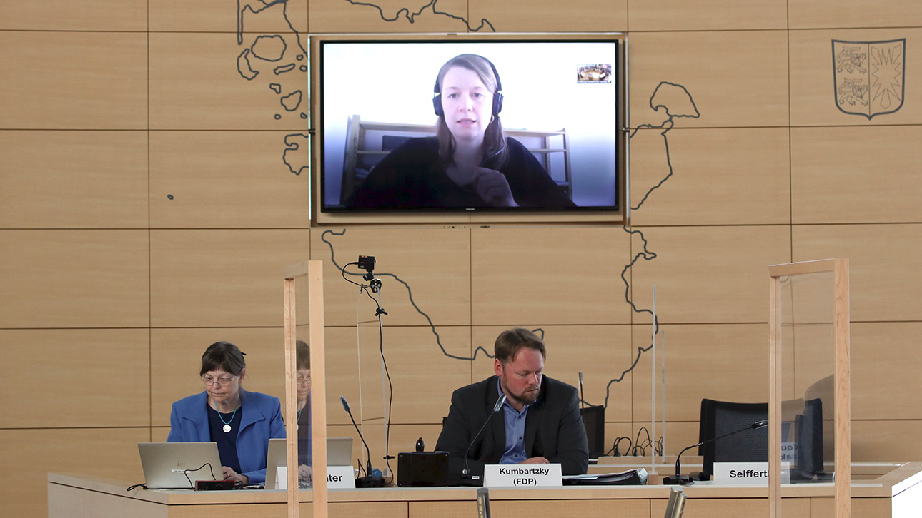 Im Plenarsaal sitzen der Vorsitzende des Umwelt- und Agrarausschusses, Oliver Kumbartzky und die Ausschussgeschäftsführerin auf den Präsidiumsplätzen. Auf einem Bildschirm ist eine Anzuhörende zugeschaltet. 