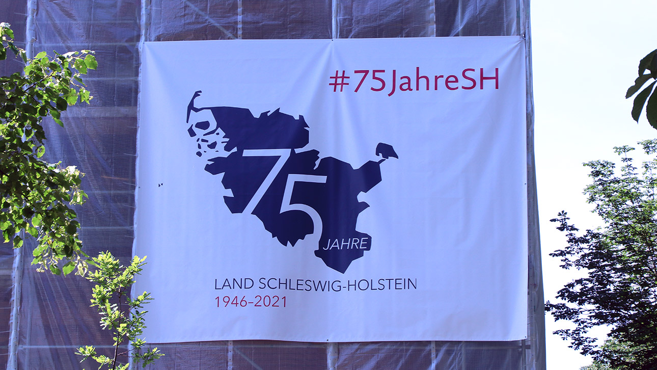 Ein weißer Banner mit einem blauen Logo für das 75. Landesjubiläum hängt an einem Baugerüst am Landeshaus.