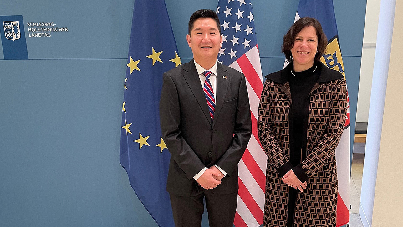 Landtagspräsidentin Kristina Herbst und Generalkonsul Jason Chue stehen im Eingangsbereich des Landeshauses vor den Flaggen der USA, Europas und Schleswig-Holsteins