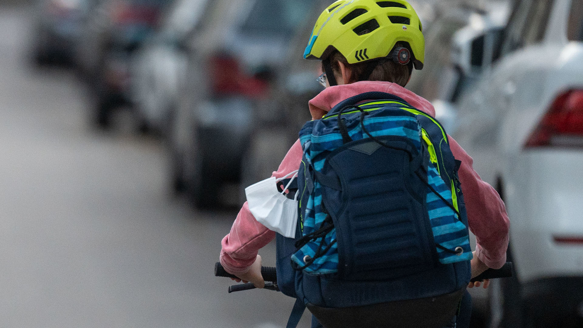 Ein Kind auf dem Fahrrad fährt auf dem Weg zur Schule auf einer Straße neben vielen geparkten Autos.