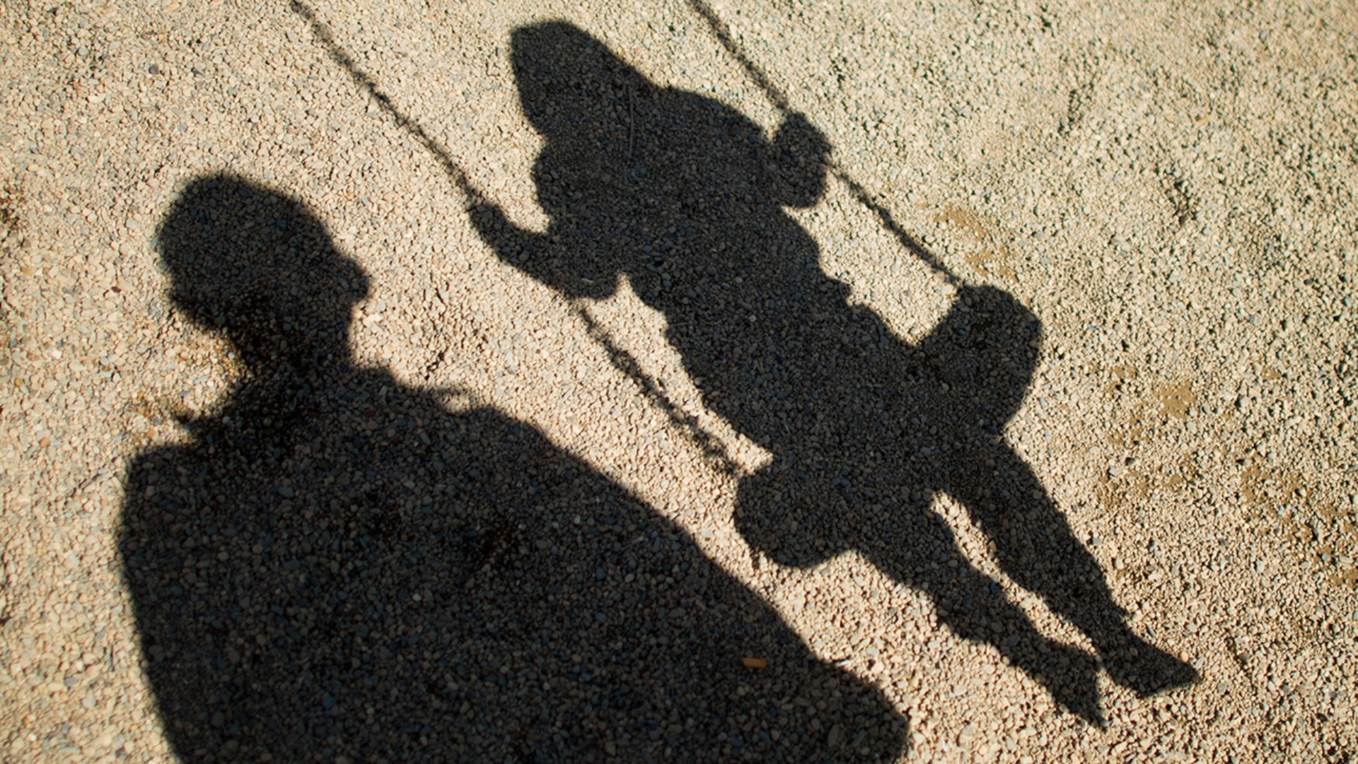 Die Schatten von einem Mann und einem schaukelnden Kind fallen auf Sand auf einem Spielplatz.
