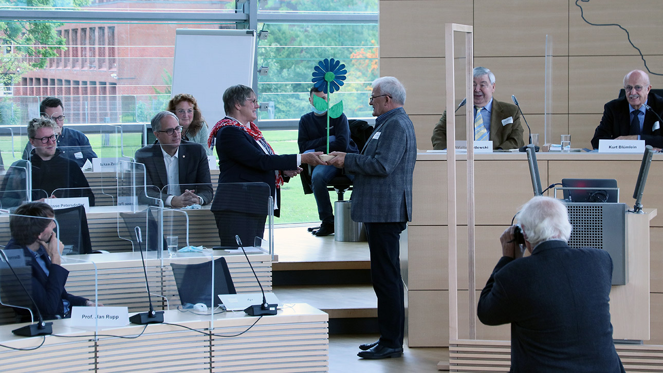 Ein Teilnehmer überreicht eine Holzblume als Geschenk an Vizepräsidentin Eickhoff-Weber.