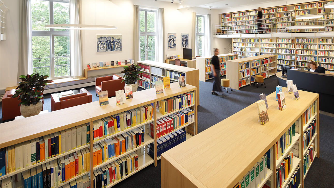Das Foto zeigt die Landtagsbibliothek mit vielen Bücherregalen, einer Leseecke und drei sich im Raum bewegenden Personen. 